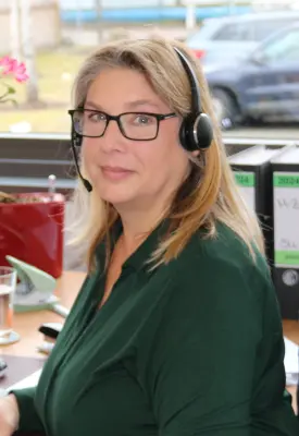 Eine blonde Frau sitzt am Arbeitsplatz. Man sieht vor Ihr eine Tastatur und einen Computerbildschirm. Im Hintergrund sieht man ein Smartphone in der Ladestation, eine Wasserflasche, ein Wasserglas und ein blühende Zimmerpflanze.
