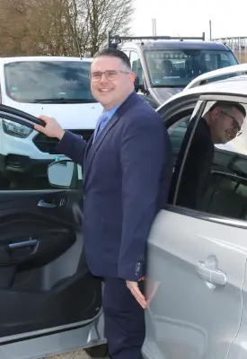 Ein Mann in den 30ern steht in der geöffneten Autotür und lächelt in die Kamera. Seine rechte Hand ruht auf der Autotür.