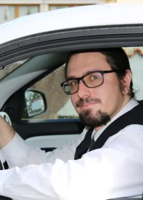 Herr S. B. schaut aus einem Autofenster, er trägt einen dunklen Anzug und weißes Hemd, er hat Kinnbart und Brille